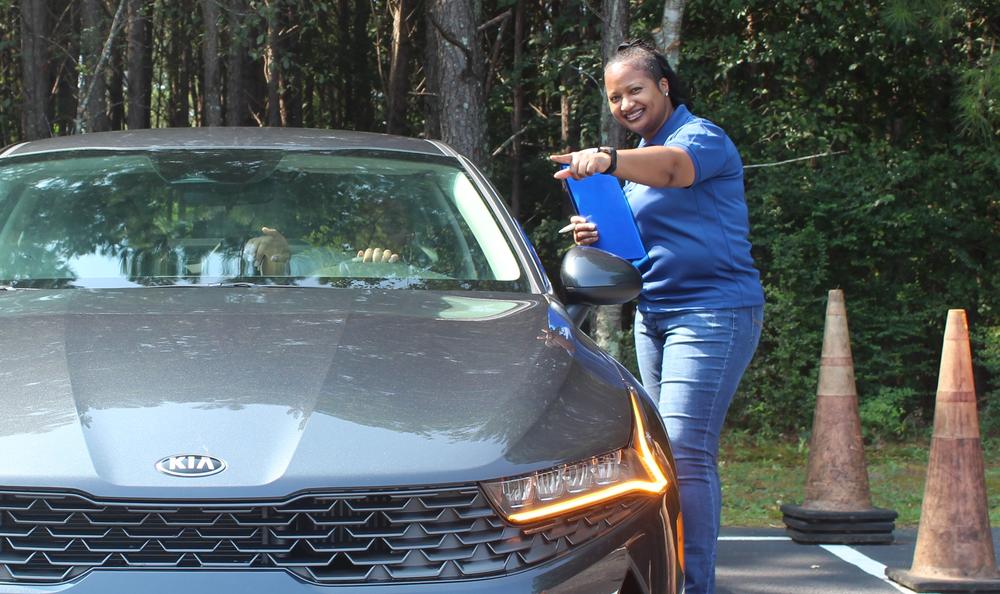 A smiling person holding a clipboard points to direct a driver forward. 