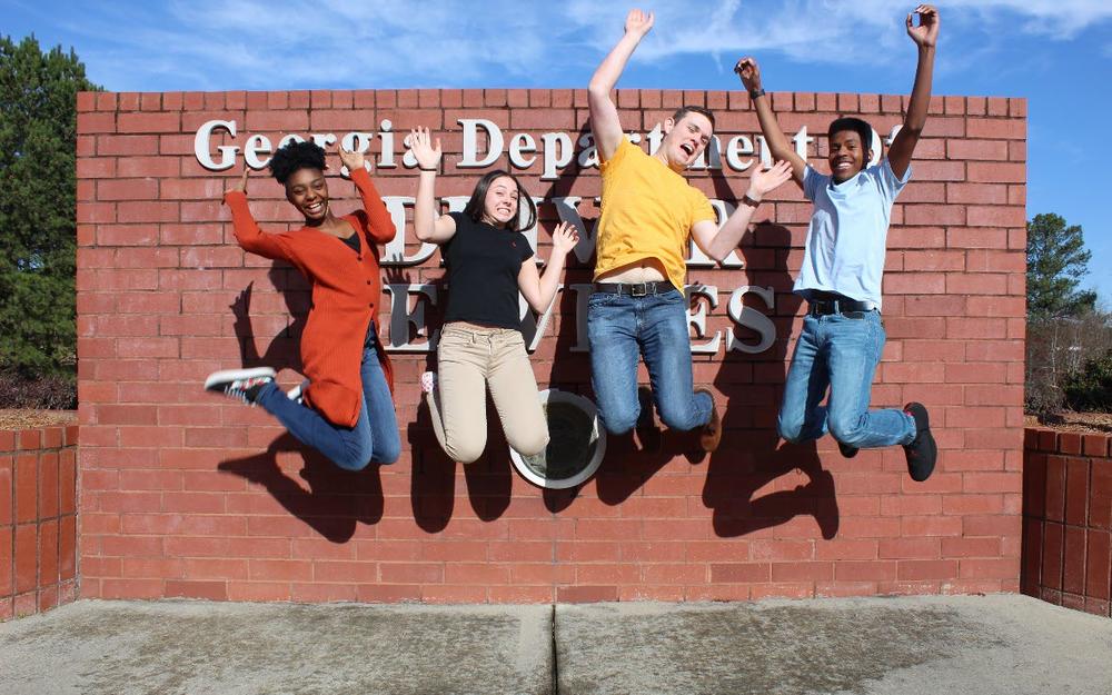 A group of four teenagers jumping for joy in front of the Georgia Department of Driver Services sign